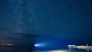 Cilvēks ar lukturīti stāv uz Latvijas jūras krasta naksnīgās, zvaigžņotās debess fonā.
