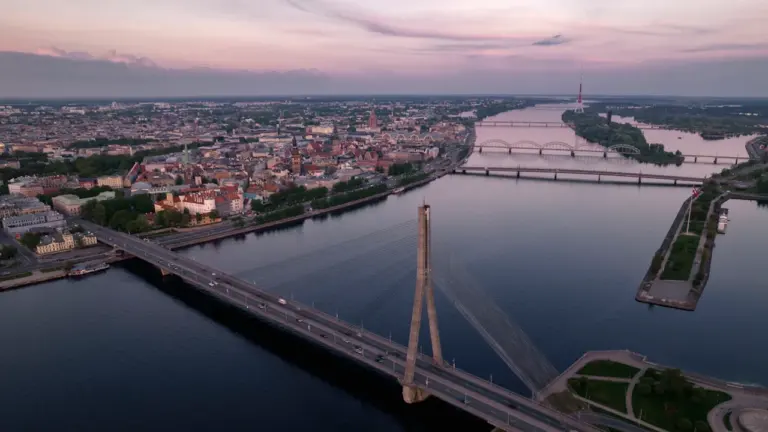 Skats uz Rīgas vēsturisko centru no putna lidojuma saulrietā, simbolizējot valsts ilgtspēju un attīstību.