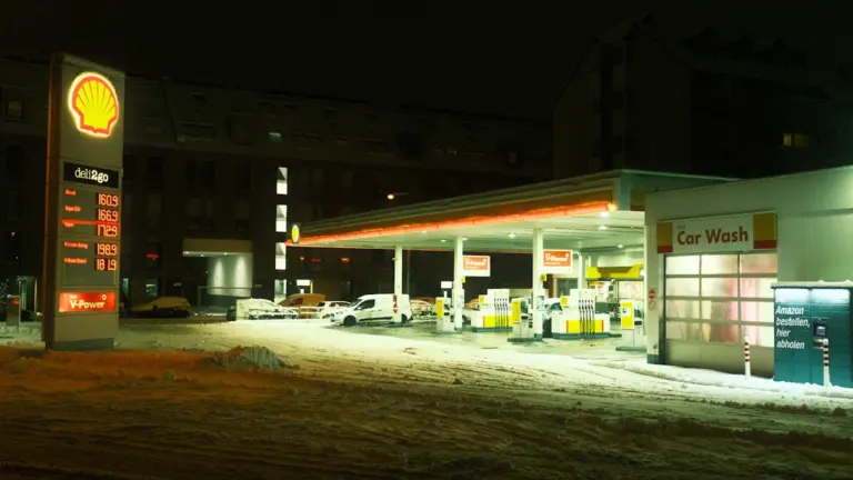 Degvielas uzpildes stacija vakarā ar sniegu, atspoguļojot Latvijas rudenīgo un ziemīgo laika apstākļu realitāti.