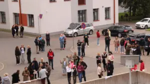 Cilvēku grupa pulcējas bruģētā laukumā pie ēkas, daudzi tur rokās papīra lapas vai kartes.