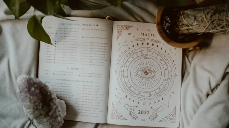 Atvērta dienasgrāmata ar zodiaka zīmju simboliem un kristālu uz gultas.